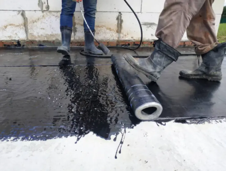 Workers applying bituminous waterproofing membrane on concrete foundation surface.